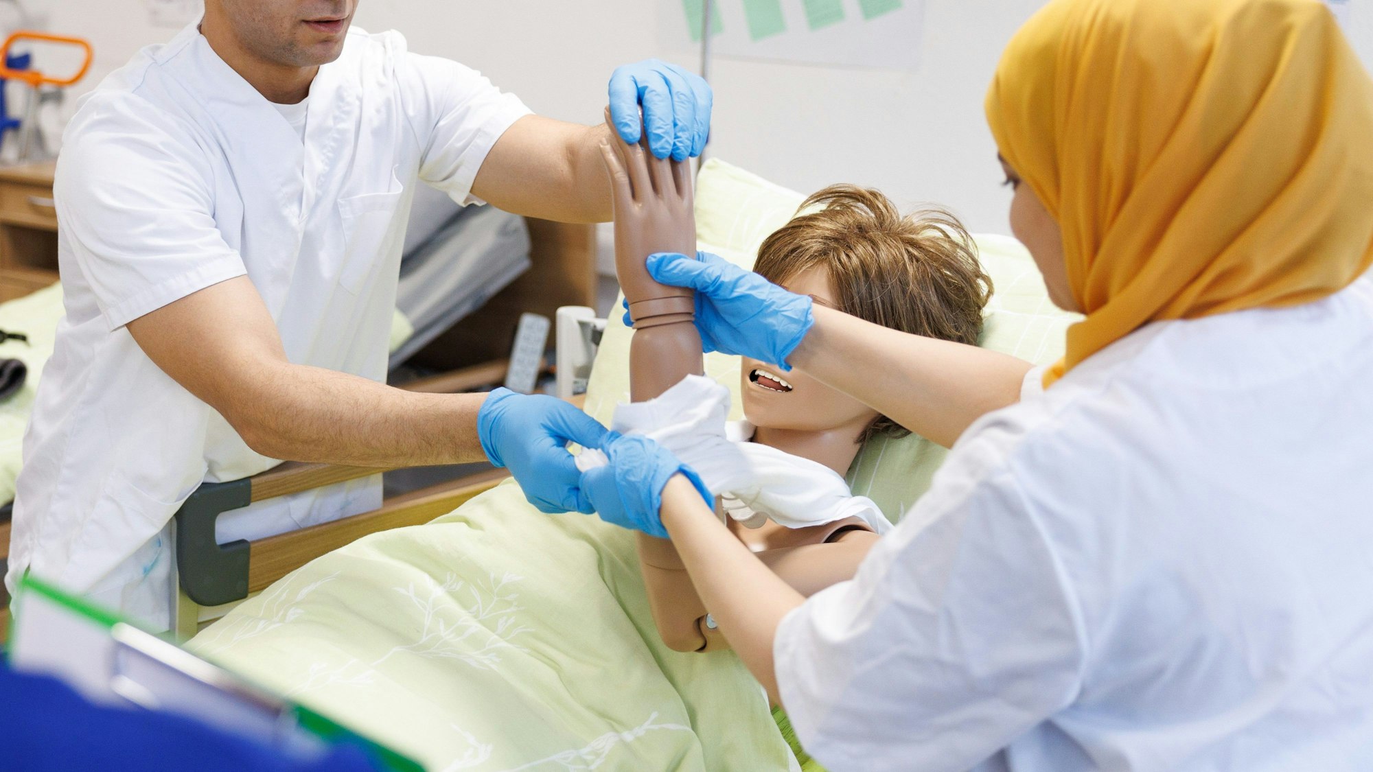 Eine Frau mit gelbem Kopftuch hält den Arm einer Körperpuppe und führt ihn durch ein T-Shirt. Ihr gegenüber steht ein Mann und assistiert ihr.
