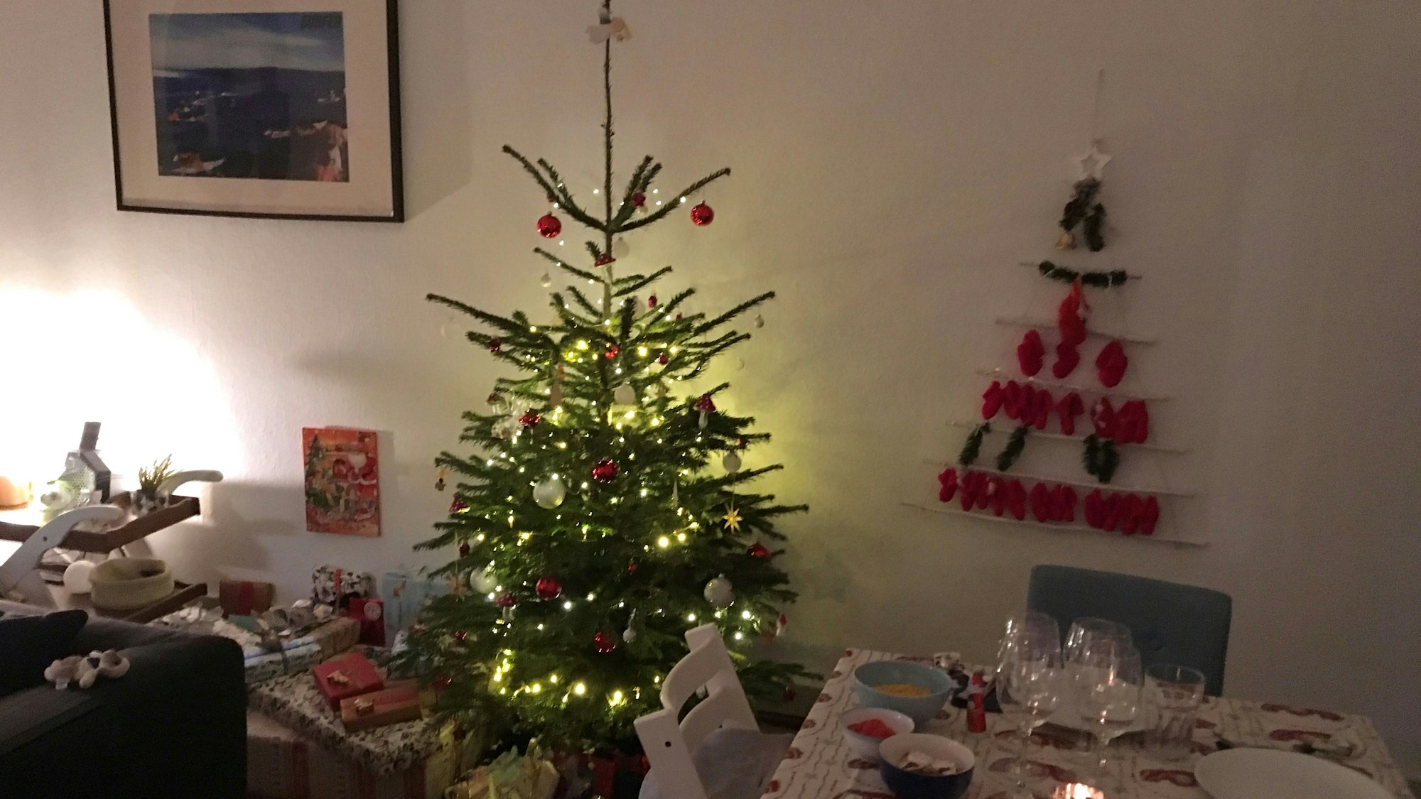 Ein rot-weiß geschmückter Weihnachtsbaum im Wohnzimmer ,daneben Geschenke und eine gedeckte Tafel.
