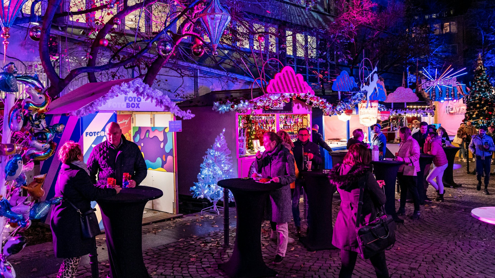 Menschen stehen an Stehtischen, pink angeleuchtete Buden im Hintergrund.