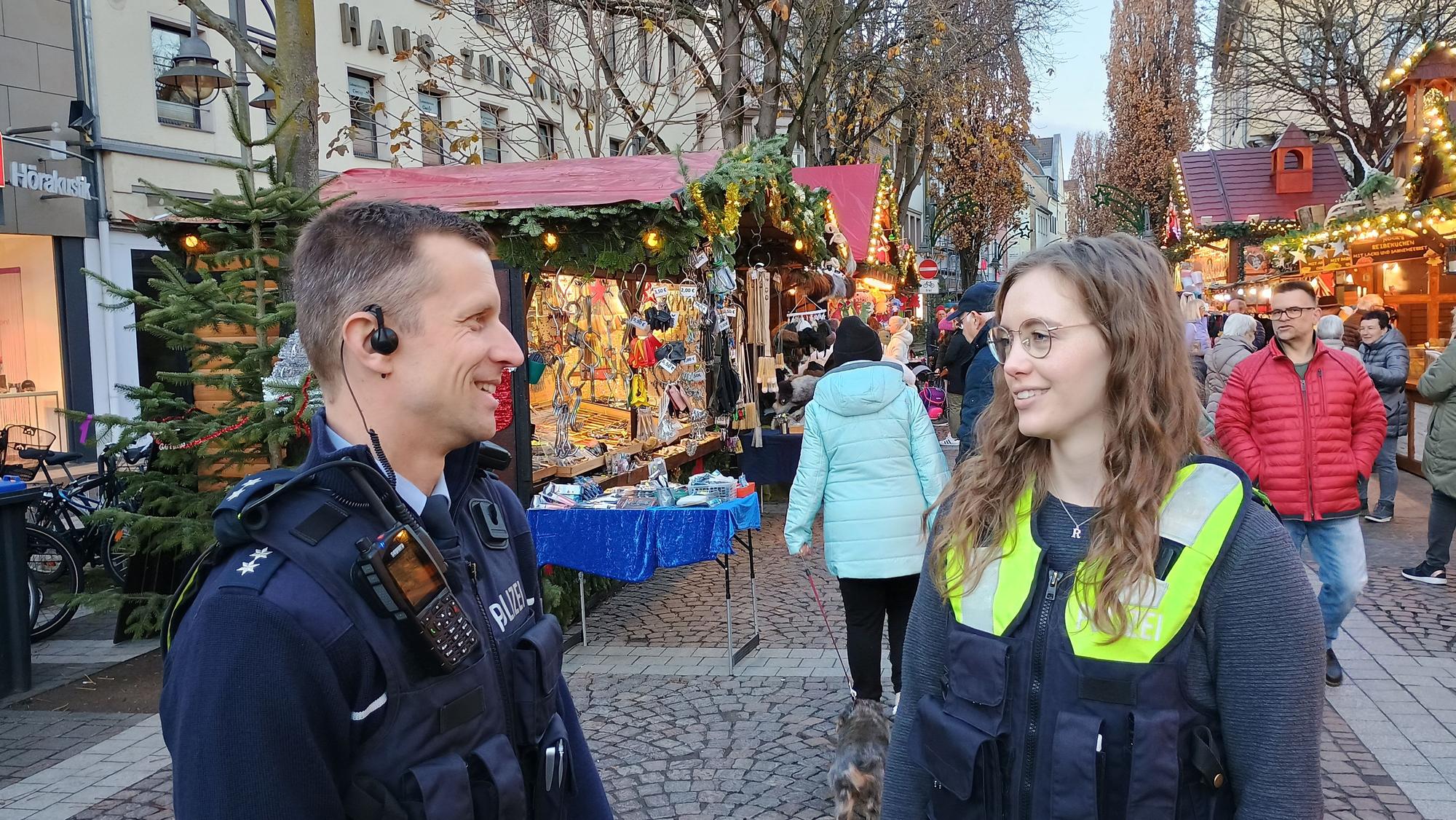 Die beiden tragen Polizeiwesten. Im Hintergrund die Stände des Weihnachtsmarktes und einige Passanten.