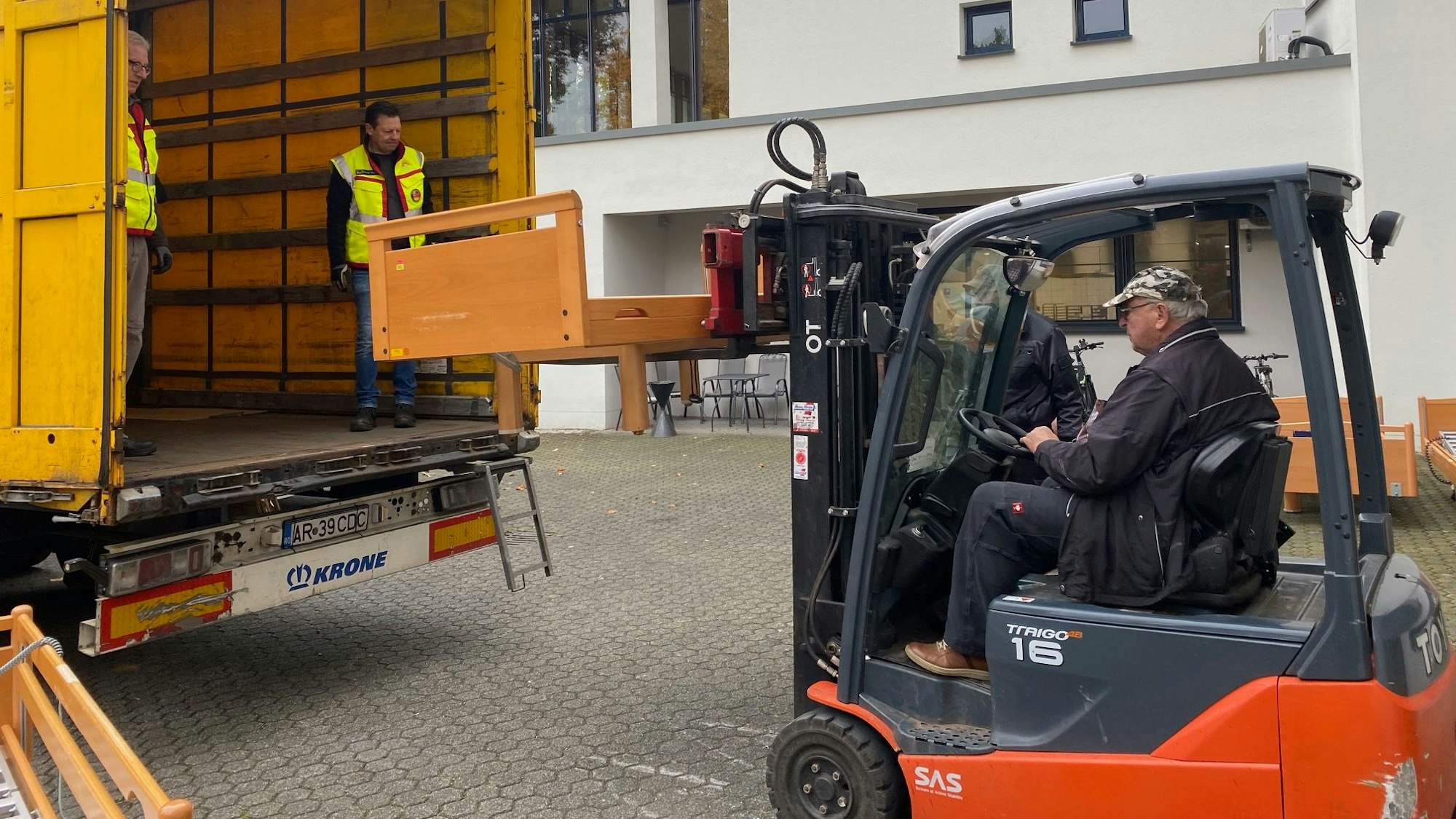 Ein Mann mit Kappe verlädt mithilfe eines roten Gabelstaplers ein Bett auf einen gelben LKW. Zwei Männer in Warnweste nehmen die Ladung entgegen.
