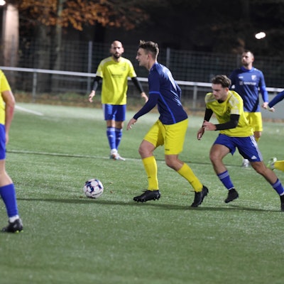 Zwei Fußballteams spielen auf einem Rasenplatz im Flutlicht abends gegeneinander.