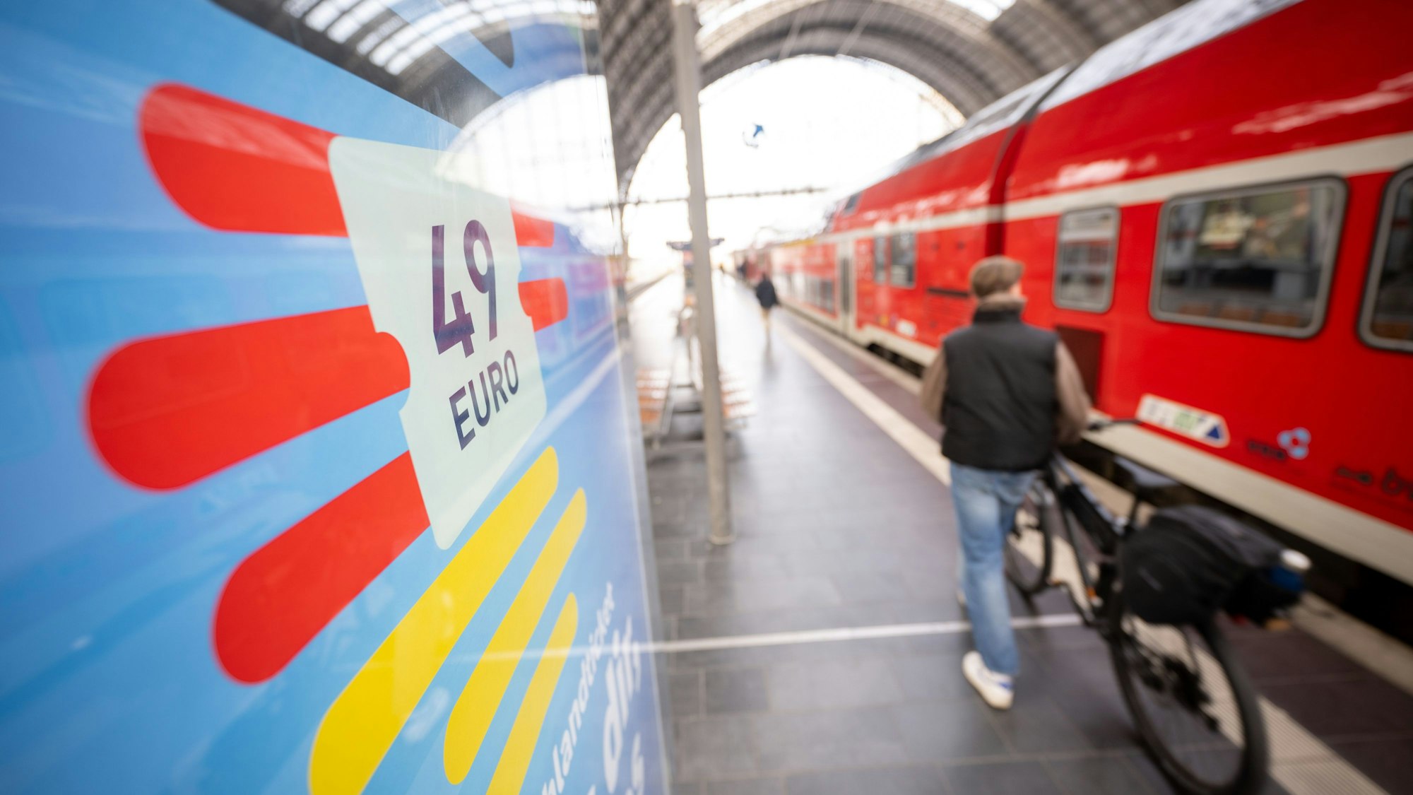 Ein Mann geht am Frankfurter Hauptbahnhof mit seinem Fahrrad zwischen der Werbung für das Deutschlandticket und einer Regionalbahn entlang.