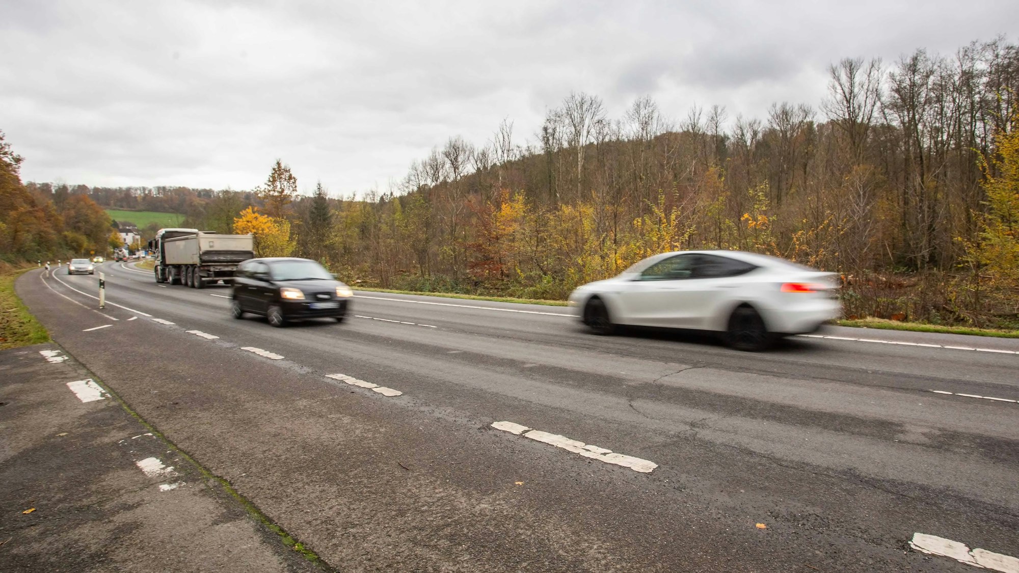 Autos fahren über eine breit ausgebaute Straße durch ein lichtes Waldgebiet.