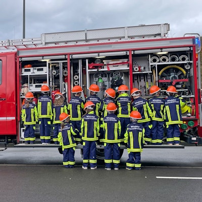 Eine Gruppe Kinder steht vor einem geöffneten Feuerwehr-Einsatzfahrzeug, mit den Rücken zum Fotografen.