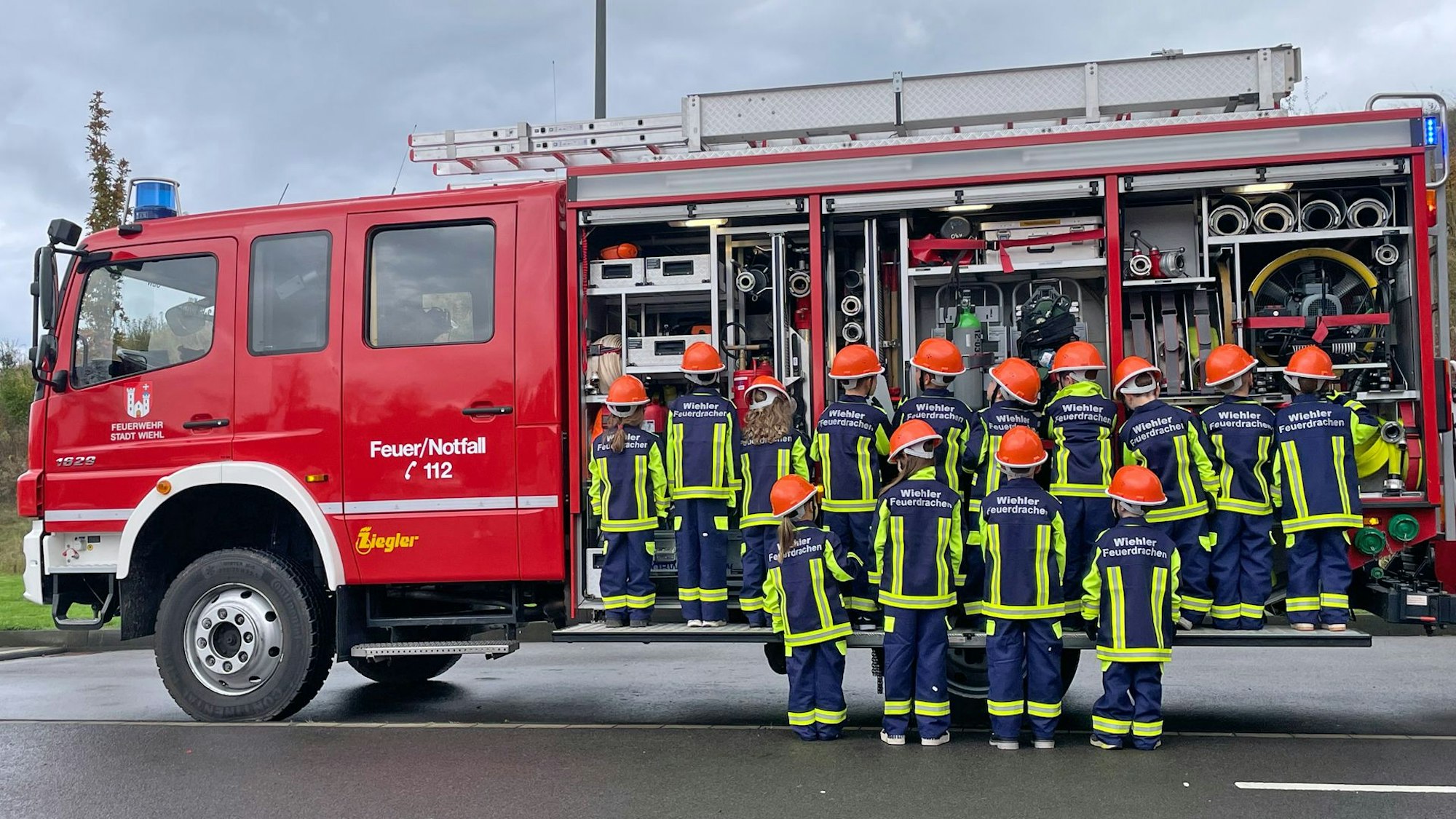Eine Gruppe Kinder steht vor einem geöffneten Feuerwehr-Einsatzfahrzeug, mit den Rücken zum Fotografen.
