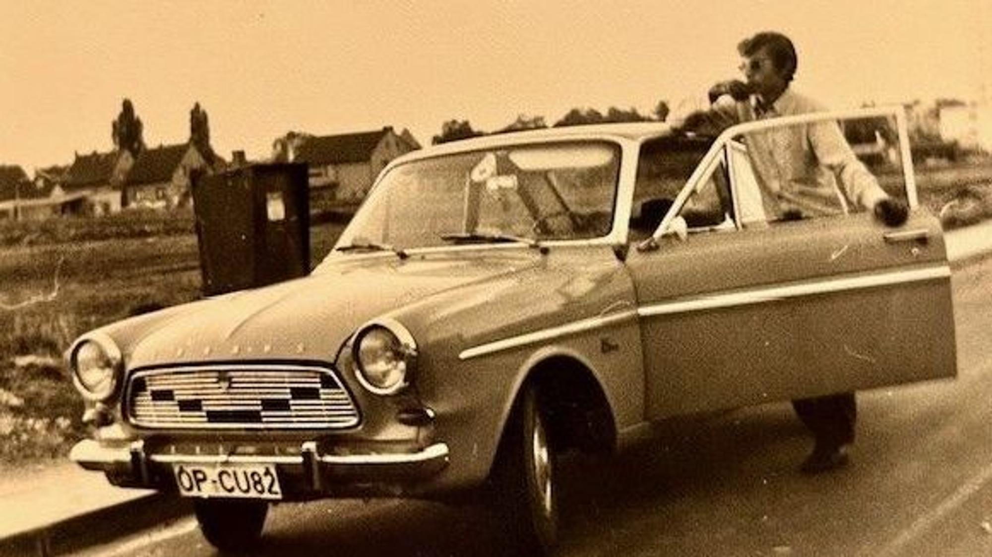 Auf dem Schwarzweißfoto ist ein Ford Taunus 12M Coupé zu sehen. In der geöffneten Fahrertür steht ein Mann und schaut über das Auto hinweg in die Landschaft.