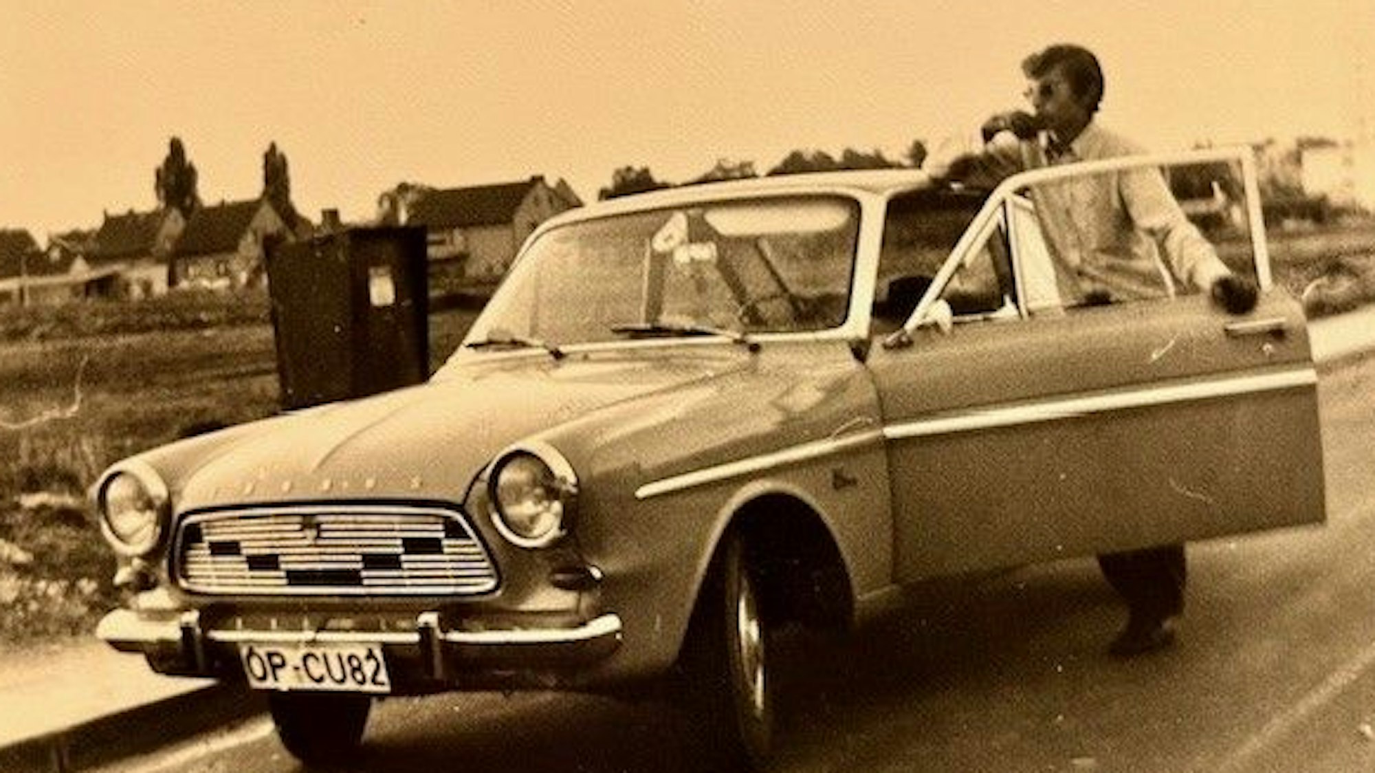 Auf dem Schwarzweißfoto ist ein Ford Taunus 12M Coupé zu sehen. In der geöffneten Fahrertür steht ein Mann und schaut über das Auto hinweg in die Landschaft.