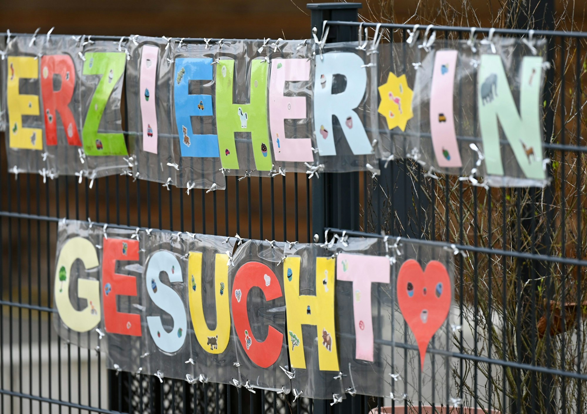 Mit bunten Buchstaben sind die Worte ·„Erzieher*in gesucht“· geformt, die an einem Zaun einer Kindertagesstätte befestigt.