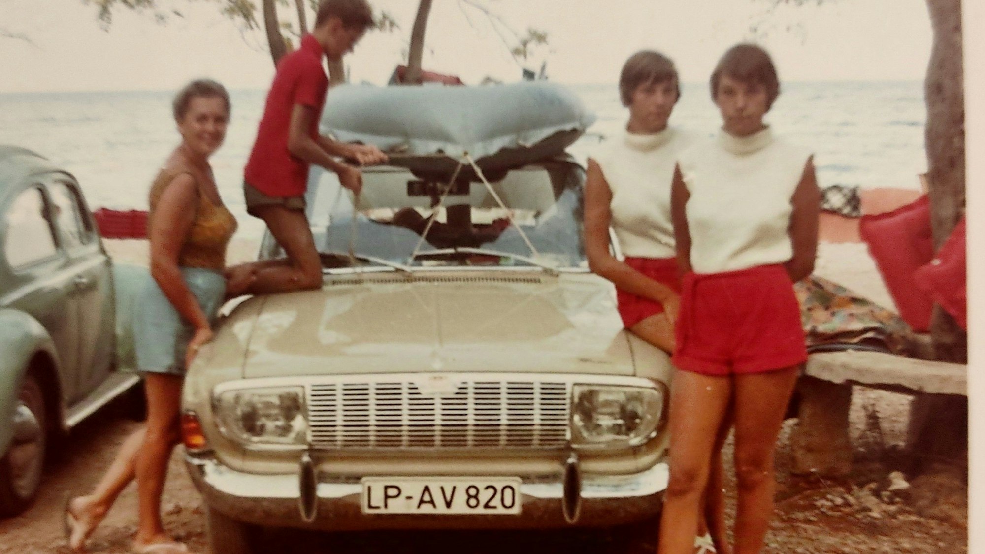 Auf einem Parkplatz am Meer gruppieren sich um einen beigen Ford mit weißem Dach zwei Mädchen im Teenager-Alter, ihre Mutter und ein jüngerer Bruder, der ein Schlauchboot auf dem Autodach befestigt.