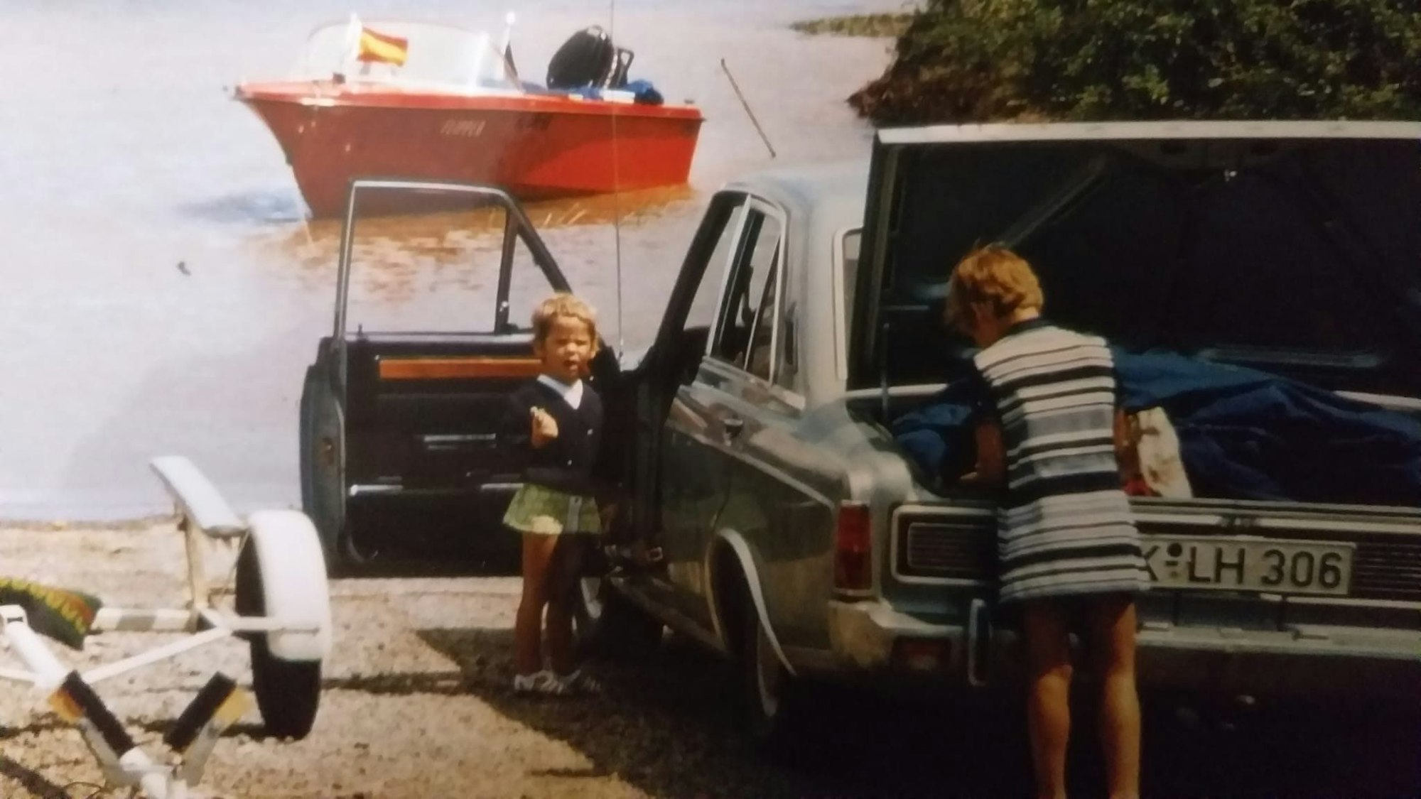 Ein grüner Ford Taunus 20M mit geöffnetem Kofferraum und geöffneter Fahrertür steht am Meeresufer. Ein Junge und ein kleines Mädchen holen Sachen aus Kofferraum und Auto.
