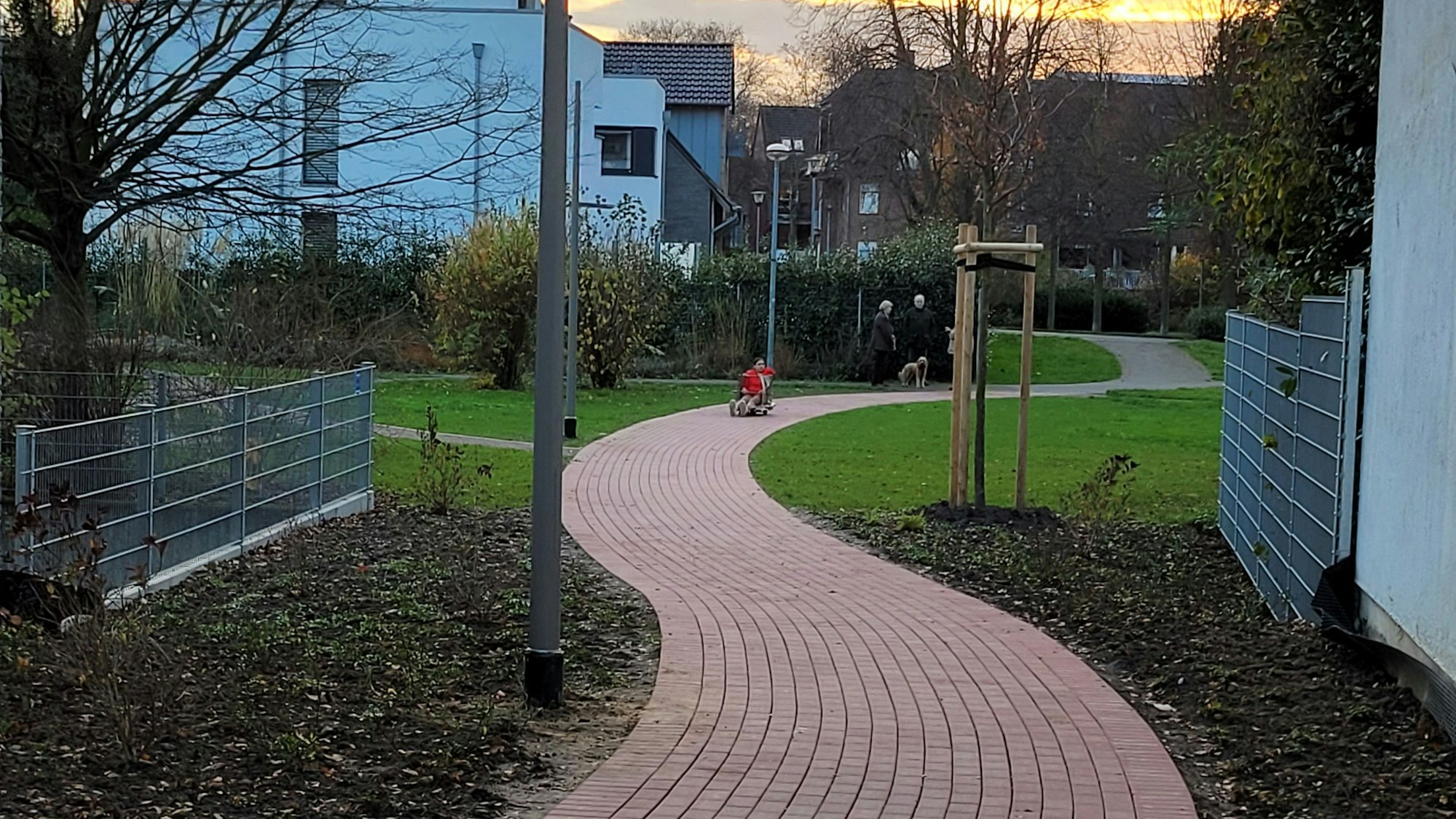 Ein geschwungener Weg, im Hintergrund links ein Gebäude und Menschen, die sich unterhalten.