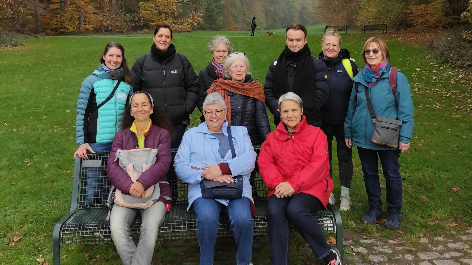 Drei Personen sitzen auf einer Parkbank, weitere Personen stehen hinter der Bank.