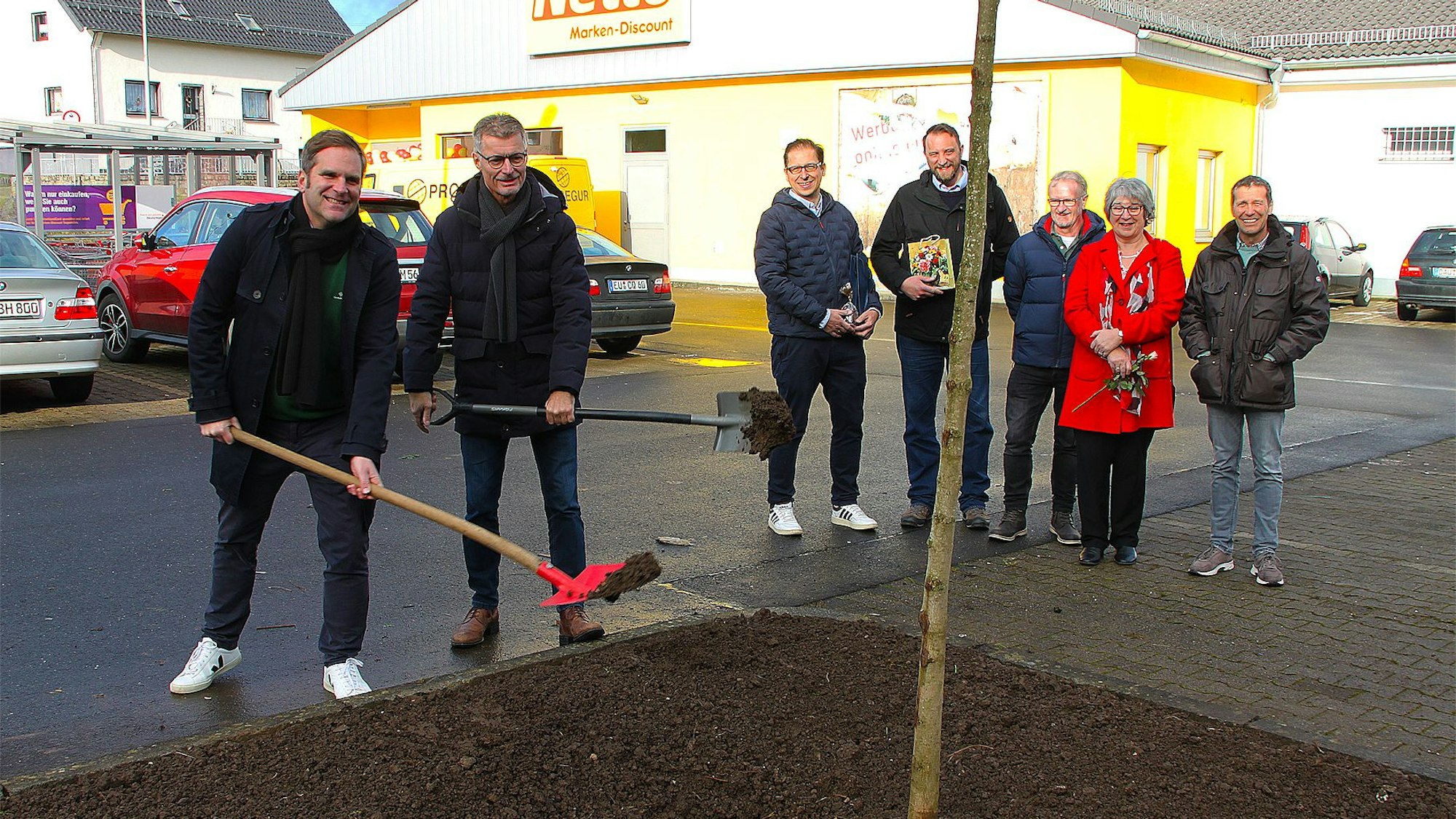 Zwei Männer schaufeln mit Spaten Erde an einen frisch gepflanzten Baum, daneben stehen einige Zuschauende. Im Hintergrund ist die Netto-Filiale zu sehen.