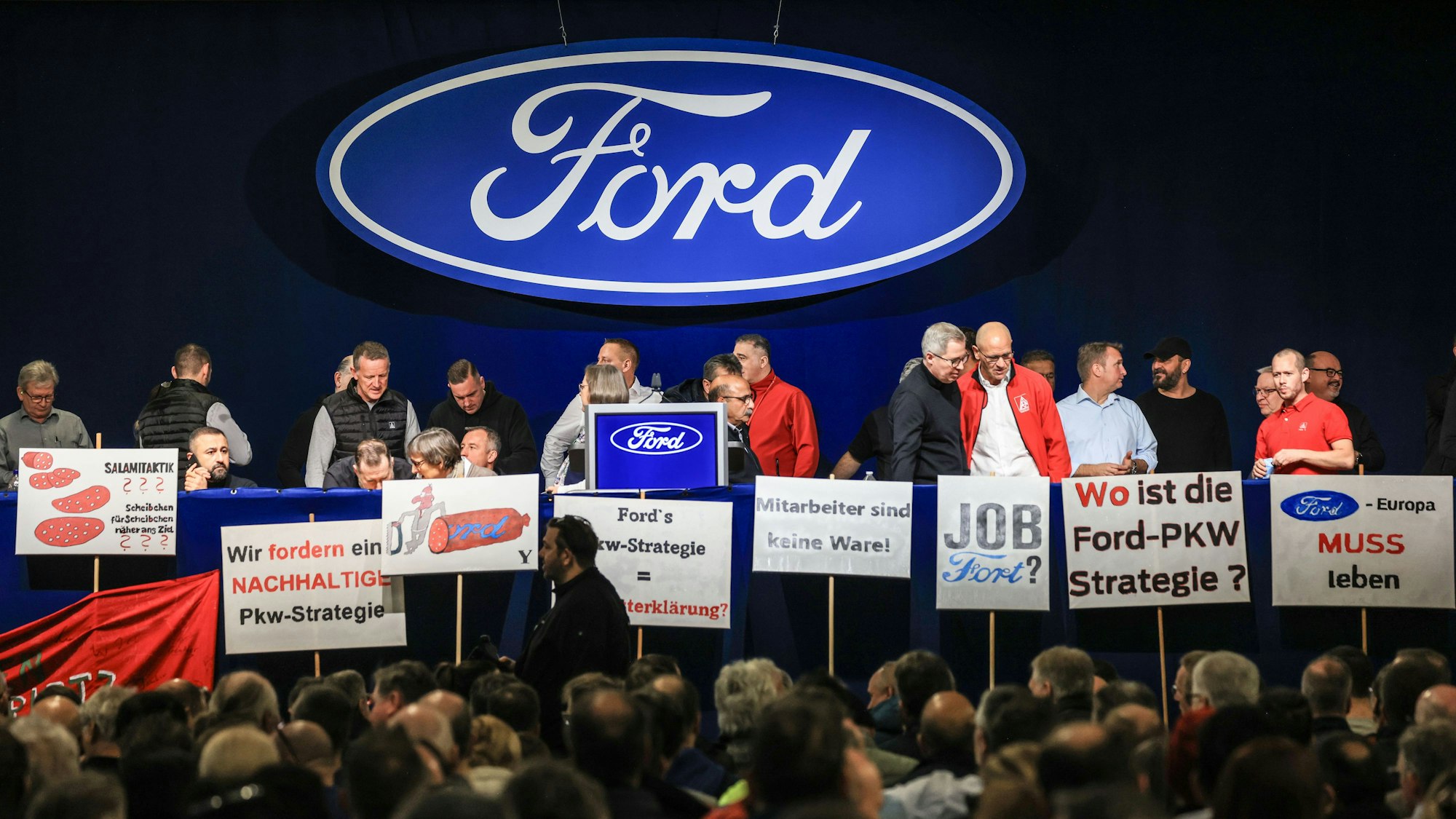 Plakate stehen vor der Bühne der Betriebsversammlung von Ford. Auf ihnen stehen Forderungen wie „Wo ist die Ford-Pkw-Strategie?“ und „Mitarbeiter sind keine Ware“. Im Bildhintergrund ist das blauweiße Firmenzeichen von Ford zu sehen.