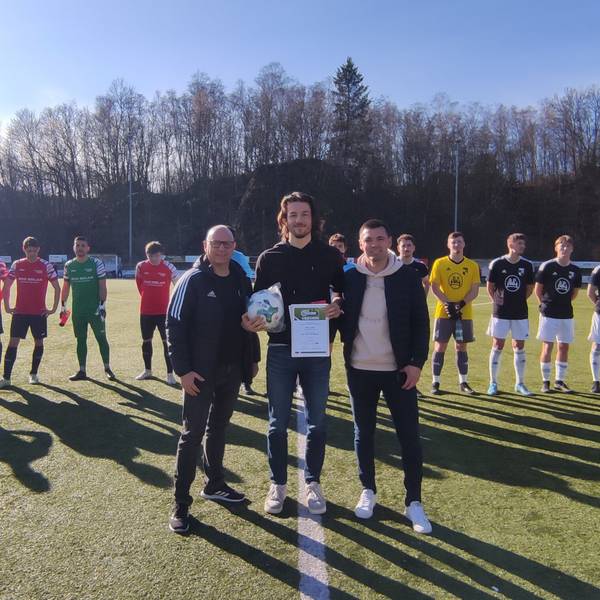 Ein Mann wird von zwei weiteren Männern auf einem Fußballplatz mit einer Urkunde ausgezeichnet. Im Hintergrund stehen zwei Mannschaften in eiern Reihe.
