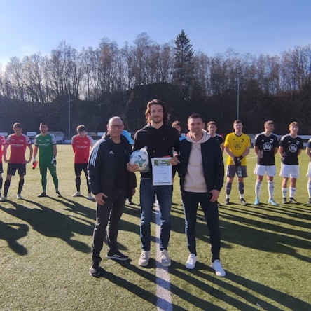 Ein Mann wird von zwei weiteren Männern auf einem Fußballplatz mit einer Urkunde ausgezeichnet. Im Hintergrund stehen zwei Mannschaften in eiern Reihe.