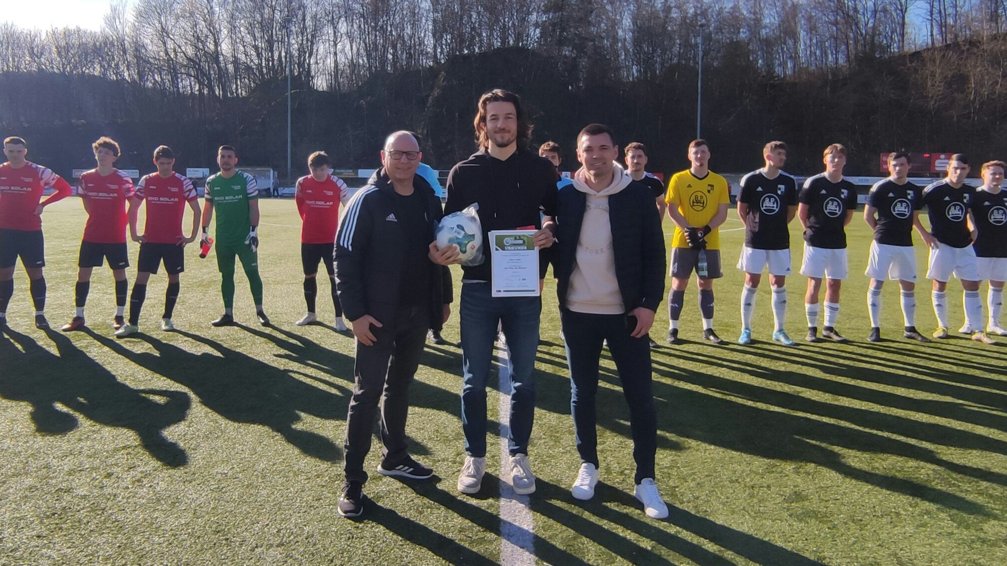 Ein Mann wird von zwei weiteren Männern auf einem Fußballplatz mit einer Urkunde ausgezeichnet. Im Hintergrund stehen zwei Mannschaften in eiern Reihe.
