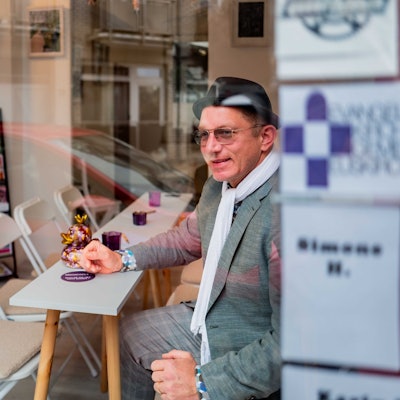 Das Bild zeigt Winfried Kubitza-Simons, der auf einem Stuhl an einem schmalen Tisch sitzt und aus dem Schaufenster schaut.