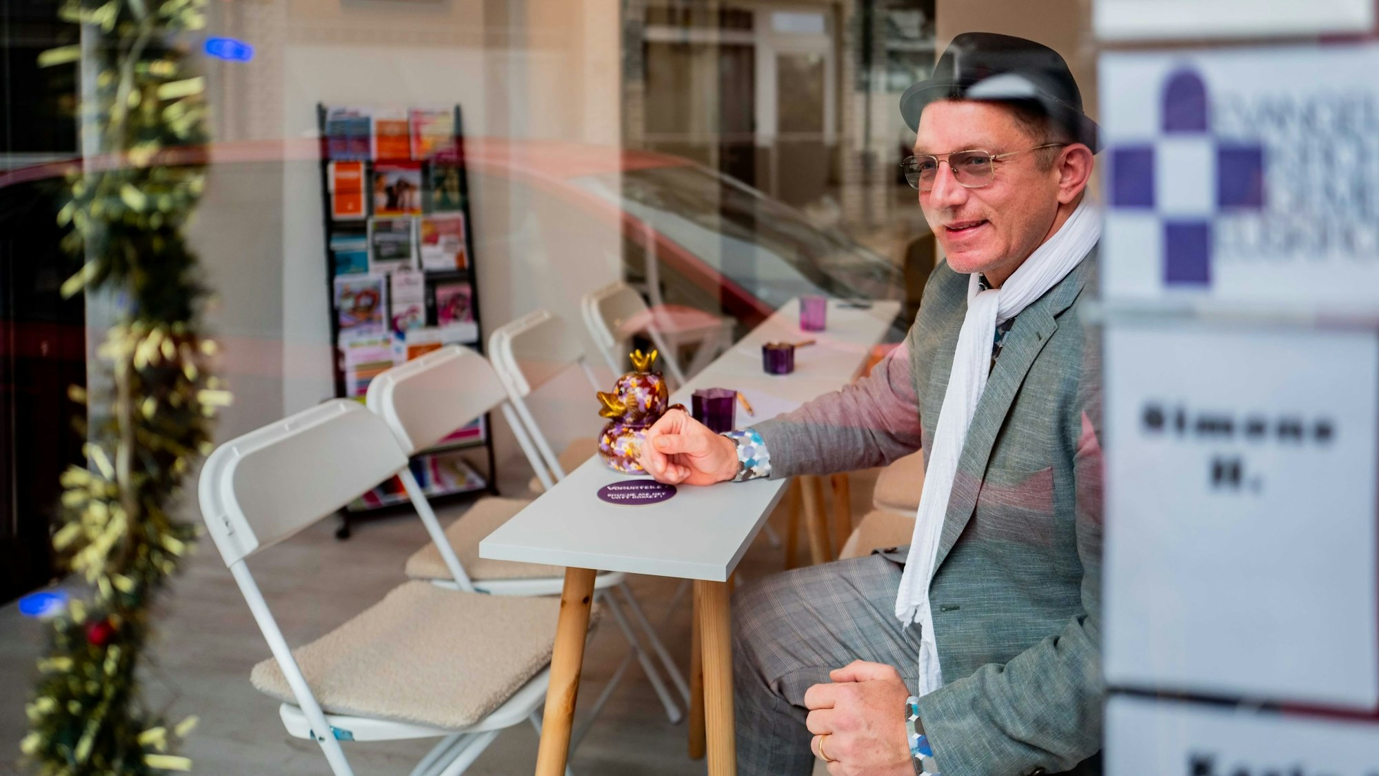 Das Bild zeigt Winfried Kubitza-Simons, der auf einem Stuhl an einem schmalen Tisch sitzt und aus dem Schaufenster schaut.
