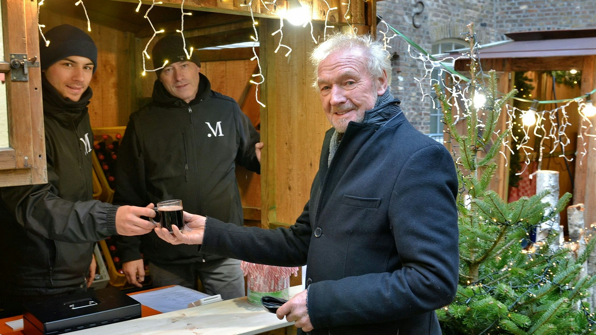 Szenerie auf einem Adventsmarkt: Ein junger Mann, der eine Mütze auf dem Kopf trägt, reicht einem älteren Herrn mit Mantel ein Glas mit einer rötlichen Flüssigkeit.