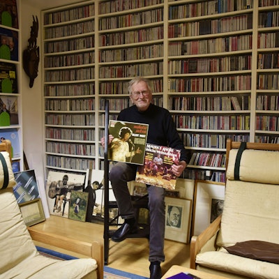 Ein älterer Herr mit Brille sitzt in einem Raum und hält zwei Plattencover in den Händen. Im Hintergrund sind haufenweise CDs in Regalen zu sehen, die bis an die Decke ragen.