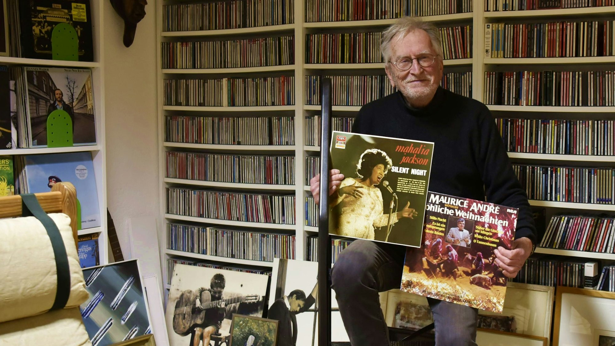 Ein älterer Herr mit Brille sitzt in einem Raum und hält zwei Plattencover in den Händen. Im Hintergrund sind haufenweise CDs in Regalen zu sehen, die bis an die Decke ragen.