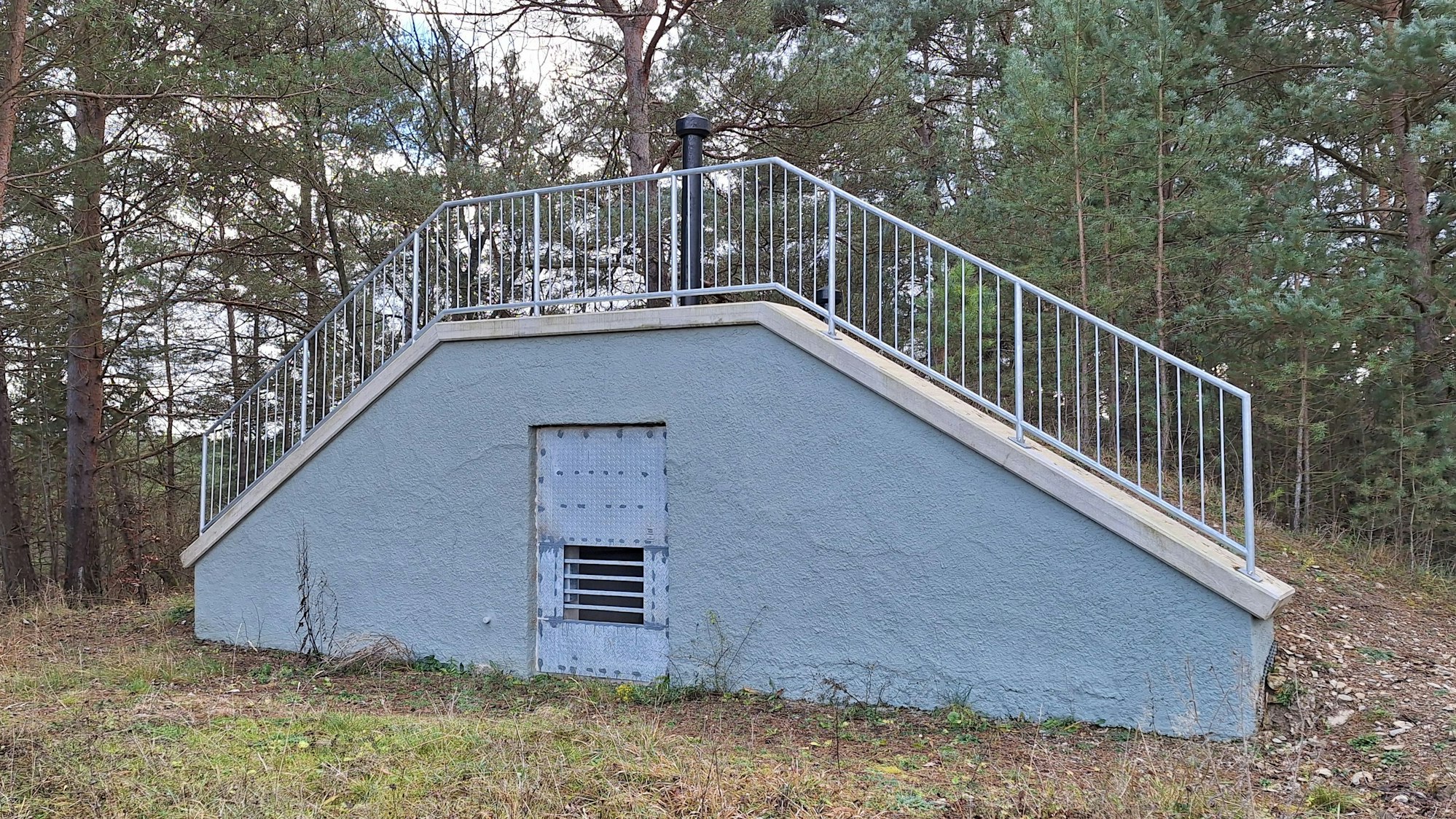 Ein trapezförmiges Gebäude, das in einem Wald steht, hat an der Forderseite eine Gittertür.
