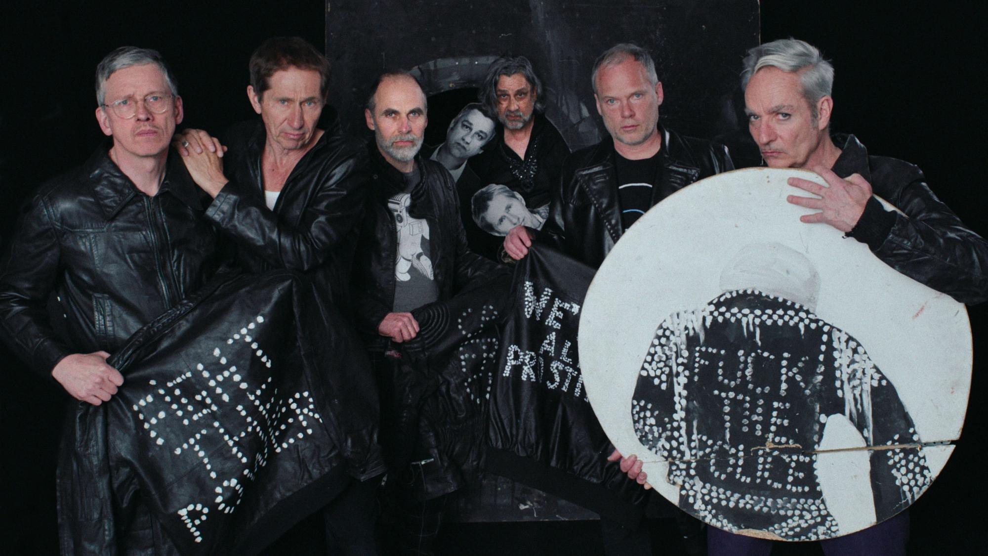 Die Goldenen Zitronen tragen schwarze Lederjacken und halten weitere Lederjacken und ein Bild mit Punk-Slogans - "Fight the Power", "Fuck the Police" - hoch.