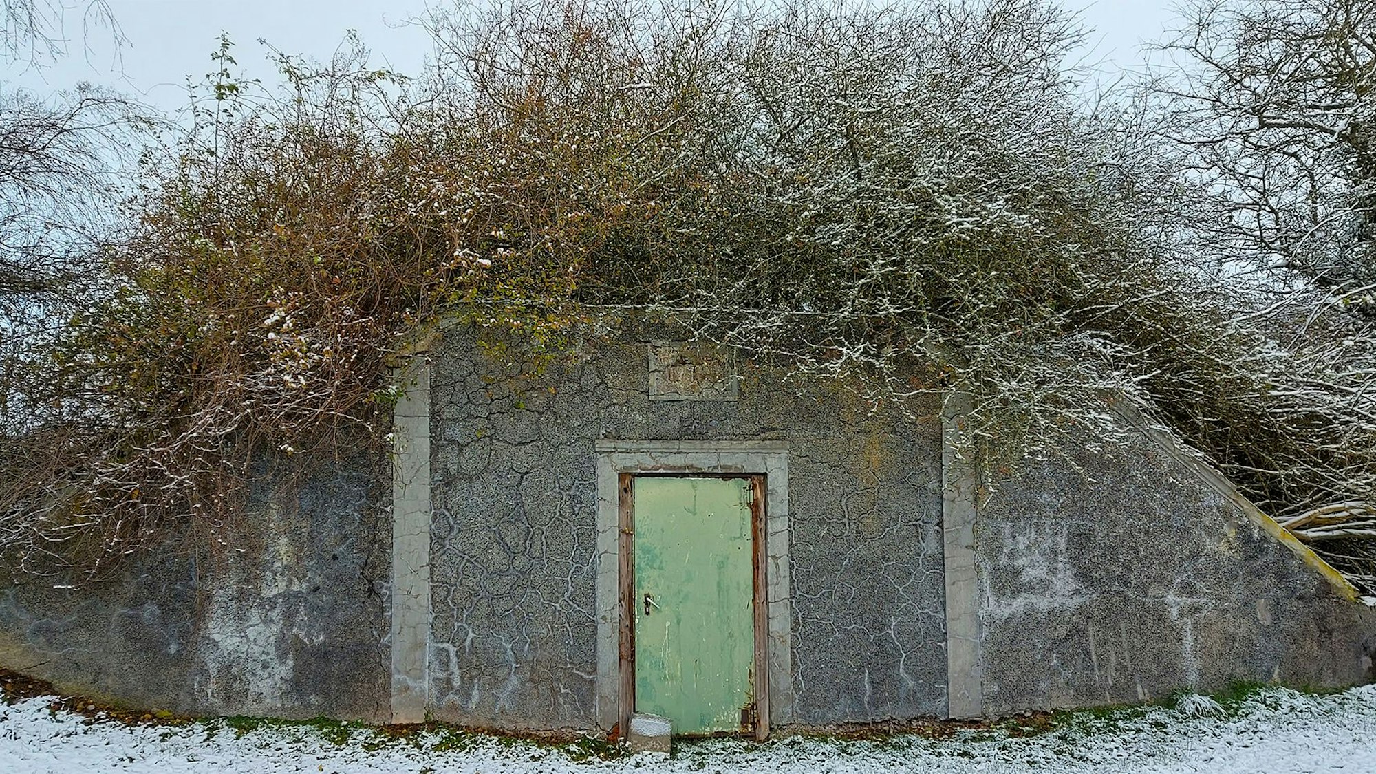 Außenansicht eines ehemaligen Wasserspeichers. Vor dem bunkerartigen Gebäude mit einer Tür liegt Schnee.