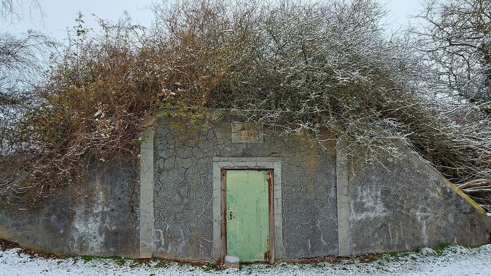 Außenansicht eines ehemaligen Wasserspeichers. Vor dem bunkerartigen Gebäude mit einer Tür liegt Schnee.