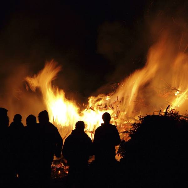 Ein großes Feuer brennt in der Nacht. Davor stehen Menschen und sehen ins Feuer.