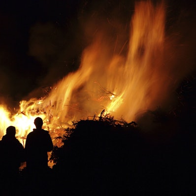 Ein großes Feuer brennt in der Nacht. Davor stehen Menschen und sehen ins Feuer.