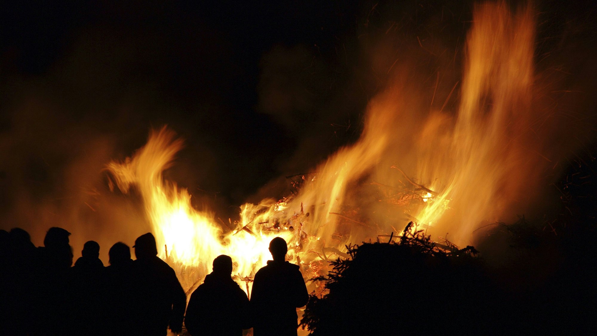 Ein großes Feuer brennt in der Nacht. Davor stehen Menschen und sehen ins Feuer.