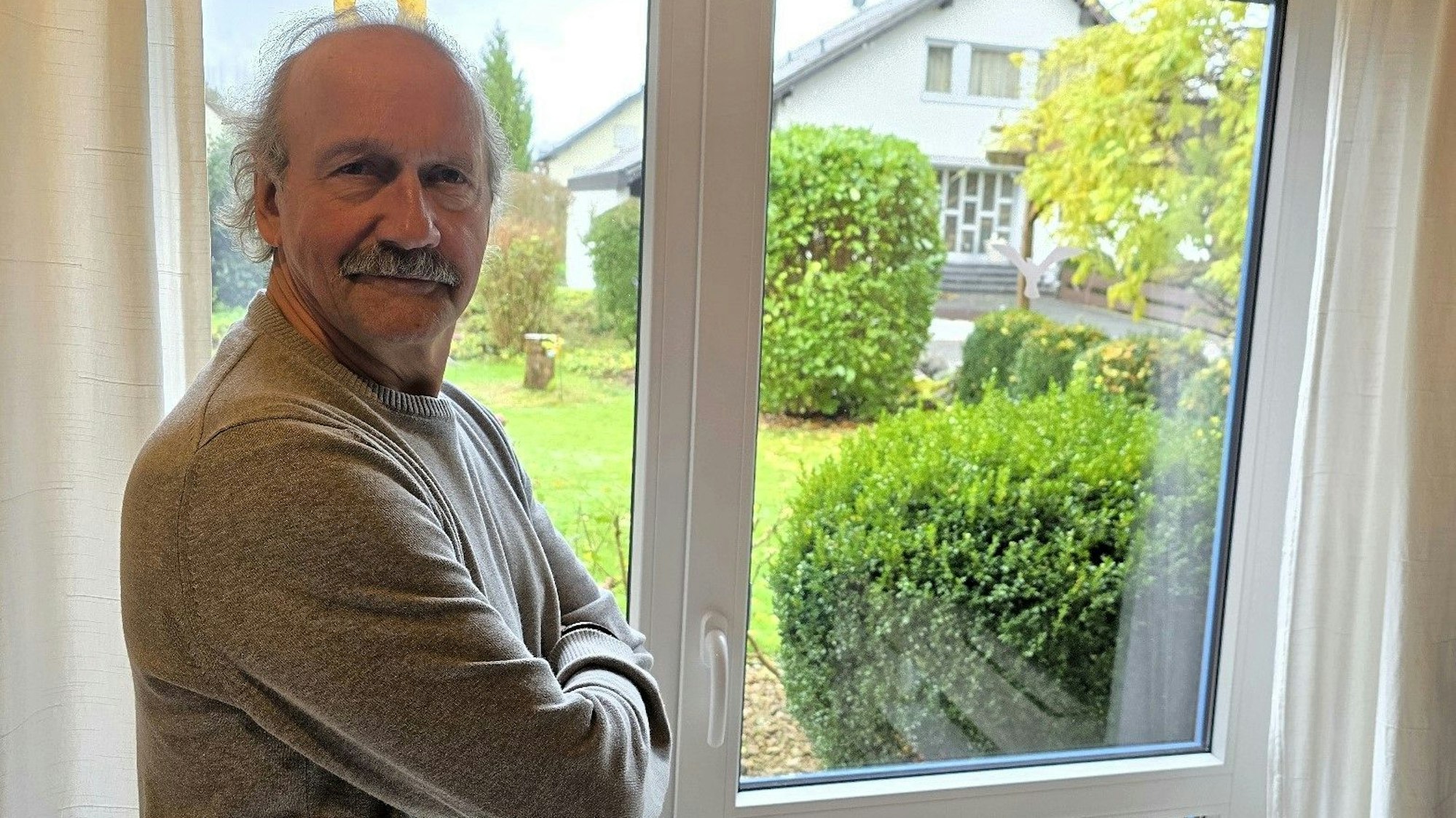 Ein älterer Herr steht mit verschränkten Armen vor einer Tür, die zu einem Garten führt, in dem Büsche stehen.