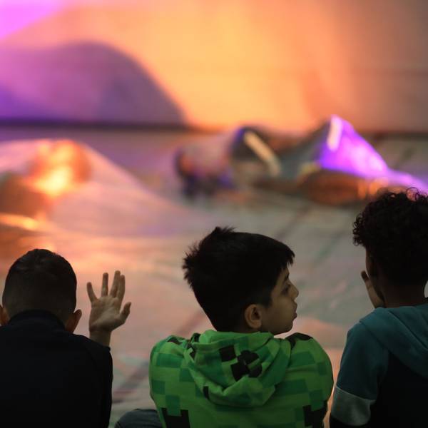 Drei Kinder sitzen auf dem Boden einer Turnhalle und schauen zwei Tänzerinnen zu, die in Seidentücher gehüllt sind.