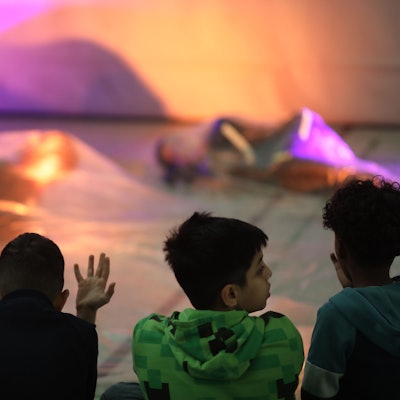 Drei Kinder sitzen auf dem Boden einer Turnhalle und schauen zwei Tänzerinnen zu, die in Seidentücher gehüllt sind.