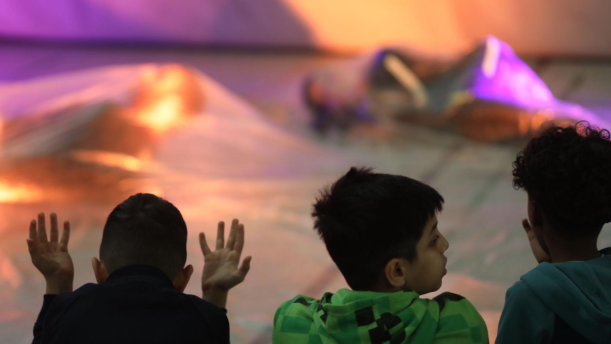Drei Kinder sitzen auf dem Boden einer Turnhalle und schauen zwei Tänzerinnen zu, die in Seidentücher gehüllt sind.