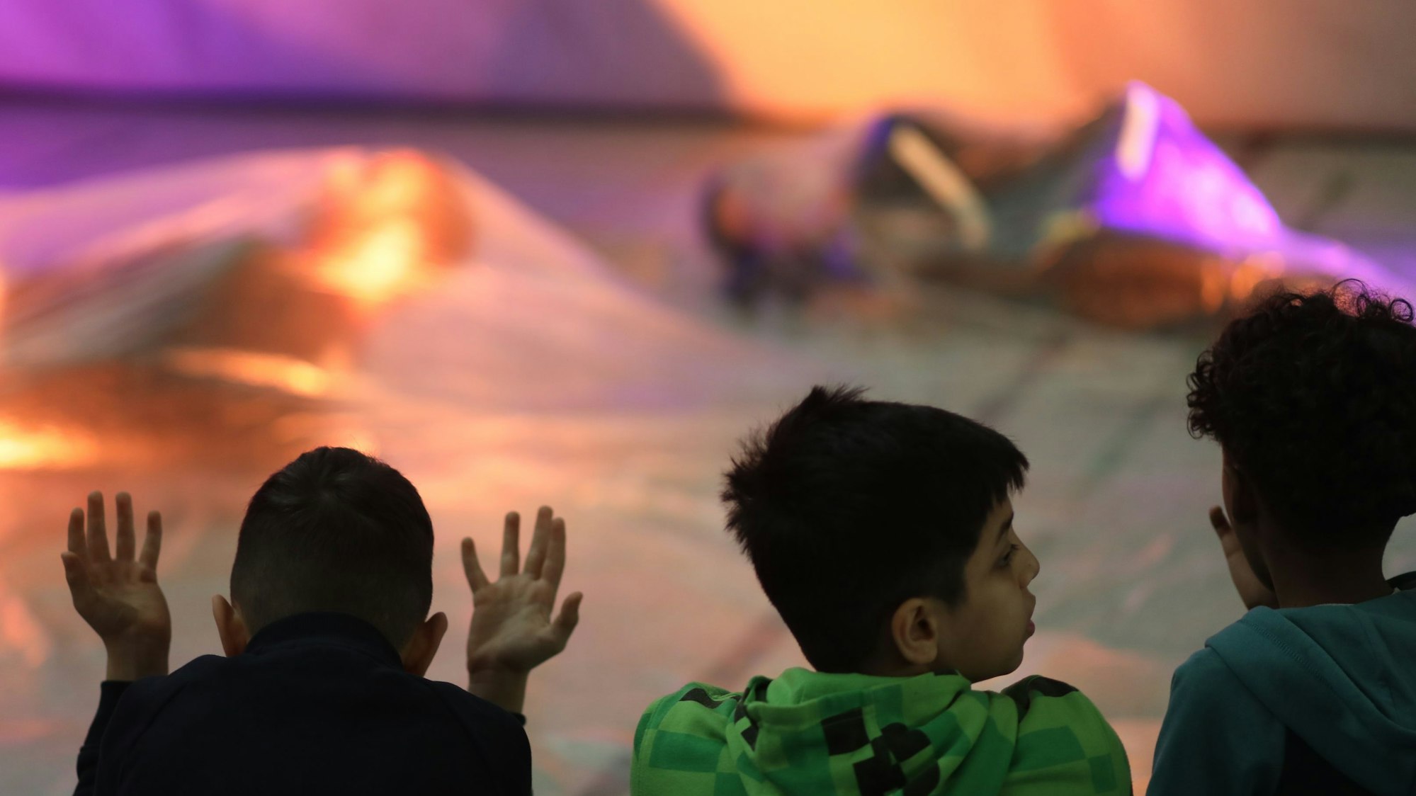 Drei Kinder sitzen auf dem Boden einer Turnhalle und schauen zwei Tänzerinnen zu, die in Seidentücher gehüllt sind.
