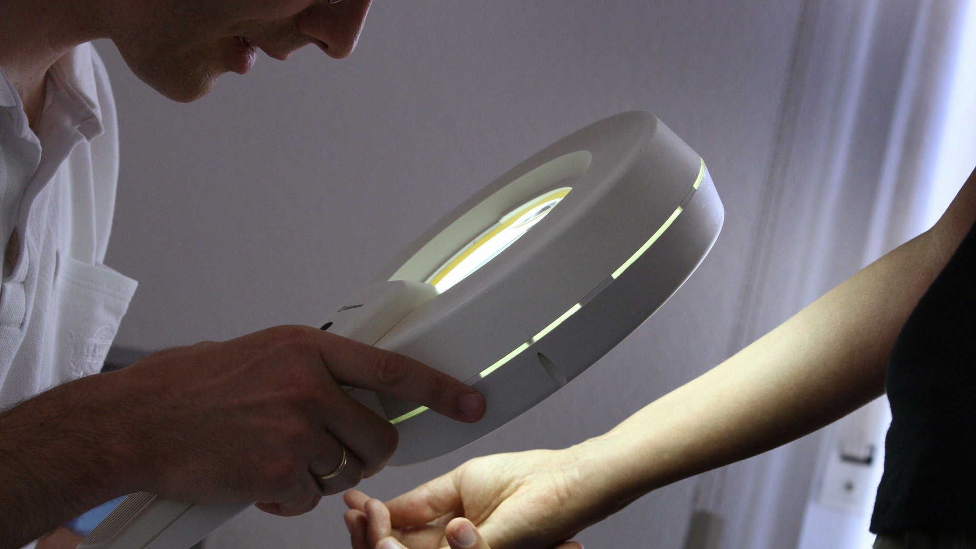 Eine Ärztin untersucht mit einer Lupe die Haut am Arm eines Patienten.