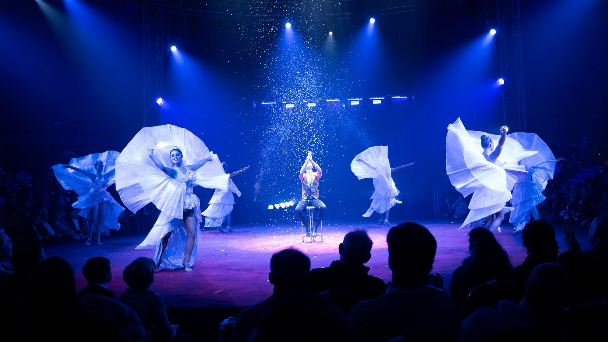 30.11.2024, Premiere des 9. Kölner WeihnachtsCircus in Köln. Eine blaubeleuchtete Manege mit fünf TänzerInnen und Konfettiregen.