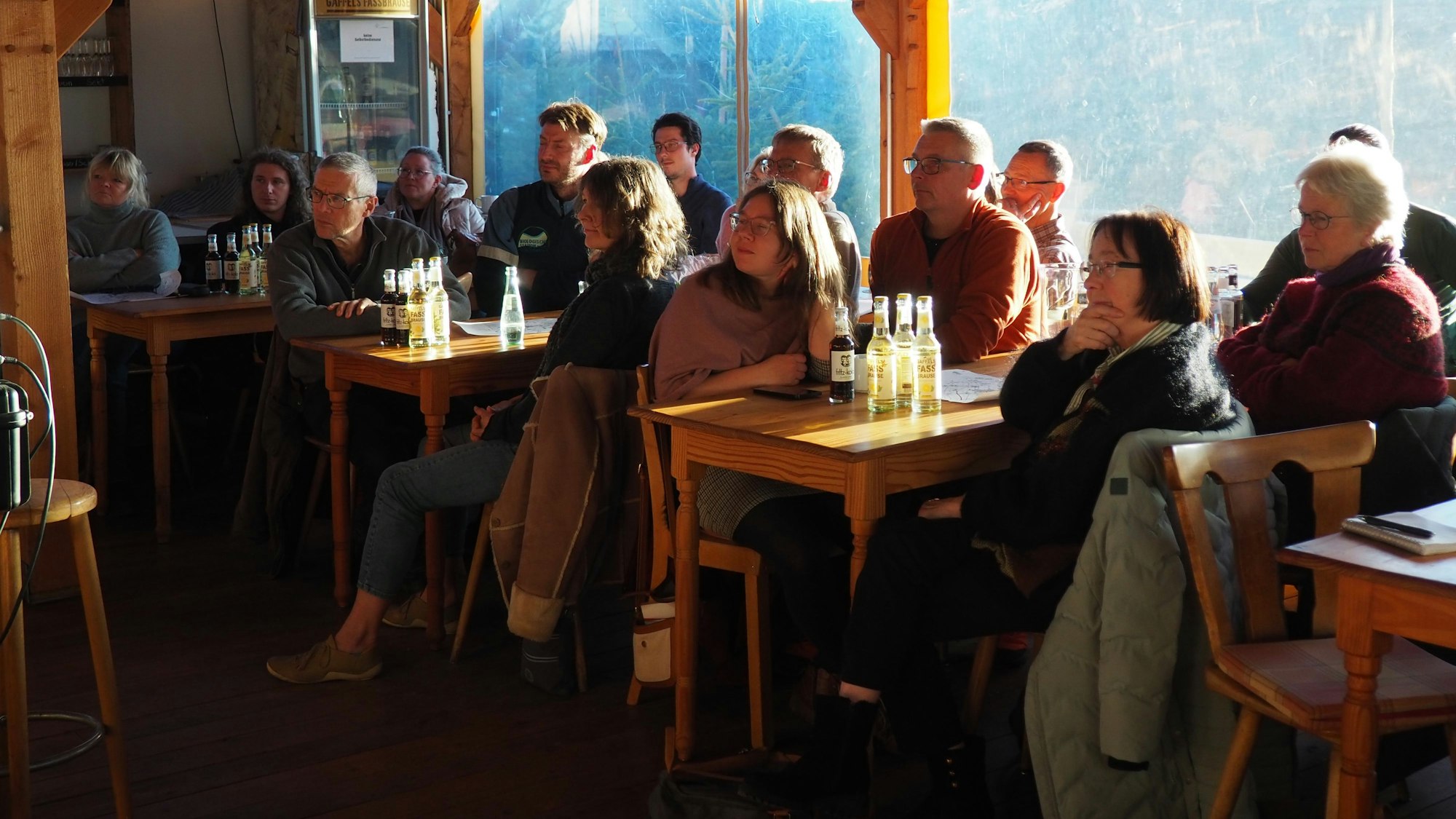 Zuhörer einer Infoveranstaltung sitzen an Tischen, auf denen Getränkeflaschen stehen.