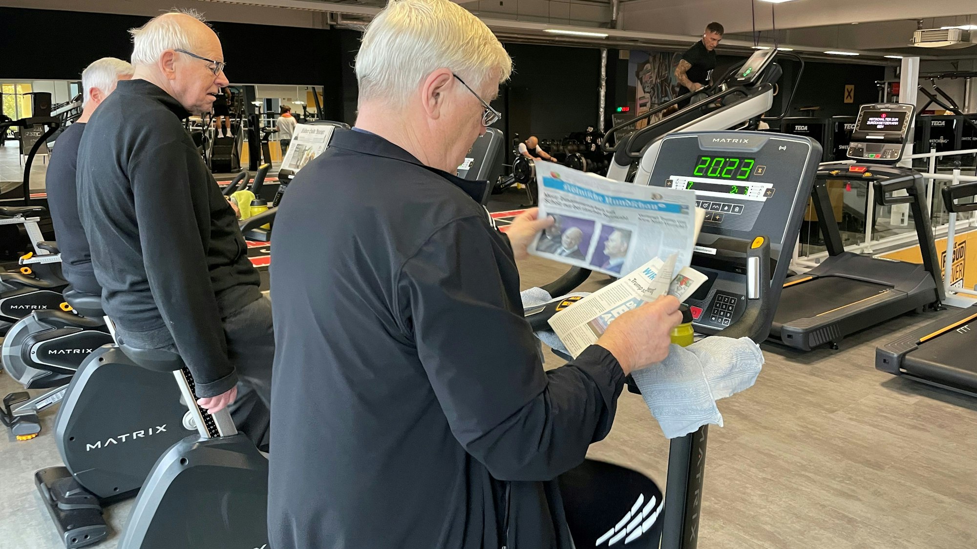 Das Bild zeigt drei Männer mit weißen Haaren, die auf dem Ergometer sitzen. Dabei lesen sie Zeitung.