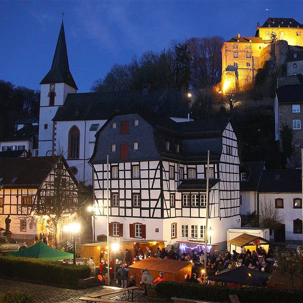 Das Bild zeigt die erleuchtete Burg in Blankenheim. Im Vordergrund ist einer kleiner Weihnachtsmarkt zu sehen.