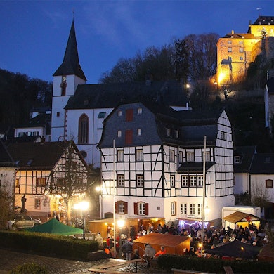 Das Bild zeigt die erleuchtete Burg in Blankenheim. Im Vordergrund ist einer kleiner Weihnachtsmarkt zu sehen.