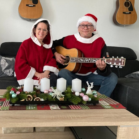Rainer Behr und Susann Behr-Lauscher sitzen in weihnachtlichen Ponchos an einem Tisch, auf dem Adventskerzen stehen. Behr hält eine Gitarre.