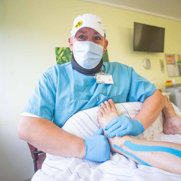 Torsten Polte trägt eine hellblaue Klinikbekleidung, eine weiße Kappe auf dem Kopf, einen Mundschutz und Handschuhe. Auf der Kappe ist ein gelbes Blindensymbol mit drei schwarzen Punkten befestigt. In einem Patientenzimmer massiert der Mann das Bein einer Patientin.