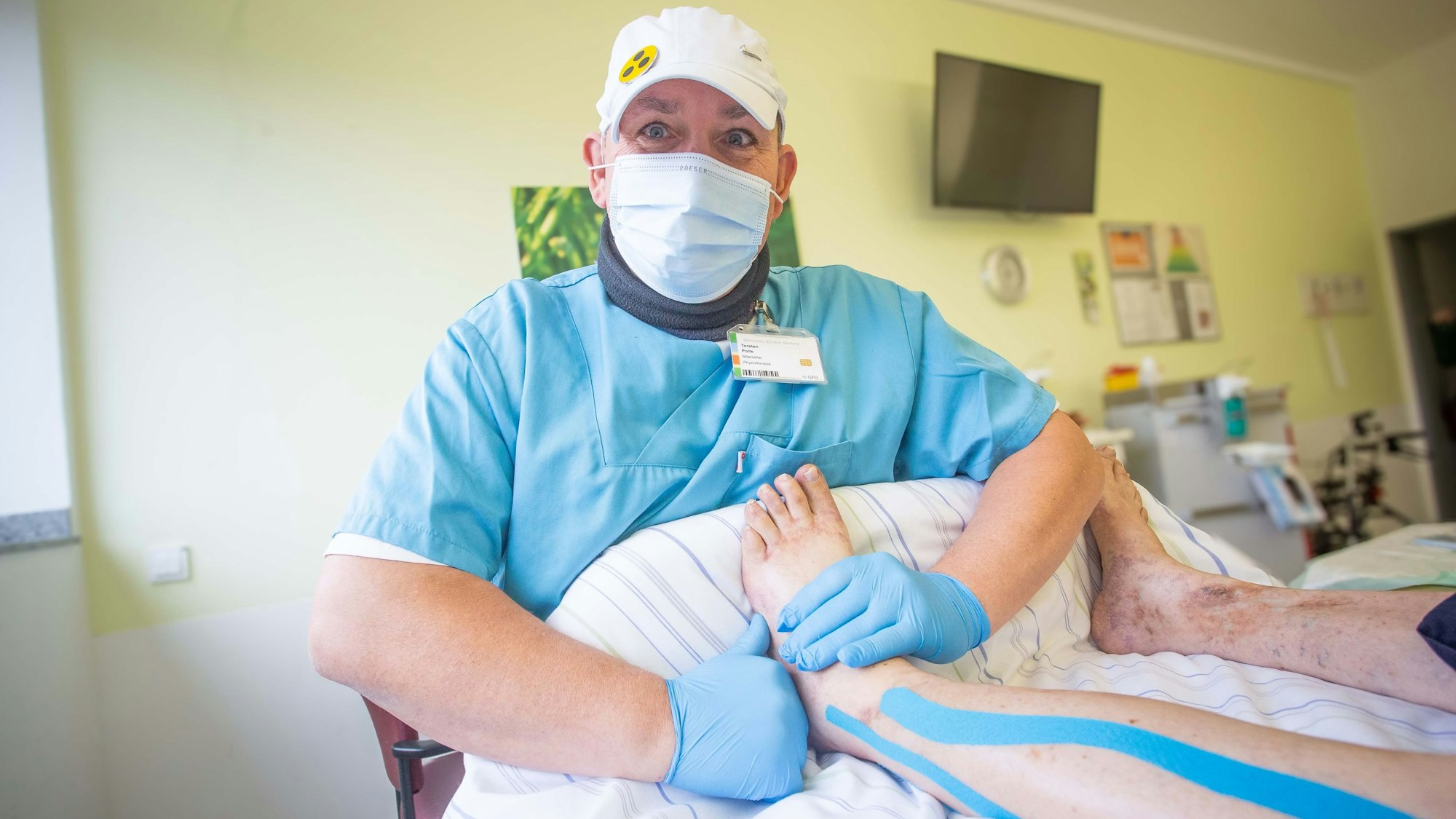 Torsten Polte trägt eine hellblaue Klinikbekleidung, eine weiße Kappe auf dem Kopf, einen Mundschutz und Handschuhe. Auf der Kappe ist ein gelbes Blindensymbol mit drei schwarzen Punkten befestigt. In einem Patientenzimmer massiert der Mann das Bein einer Patientin.