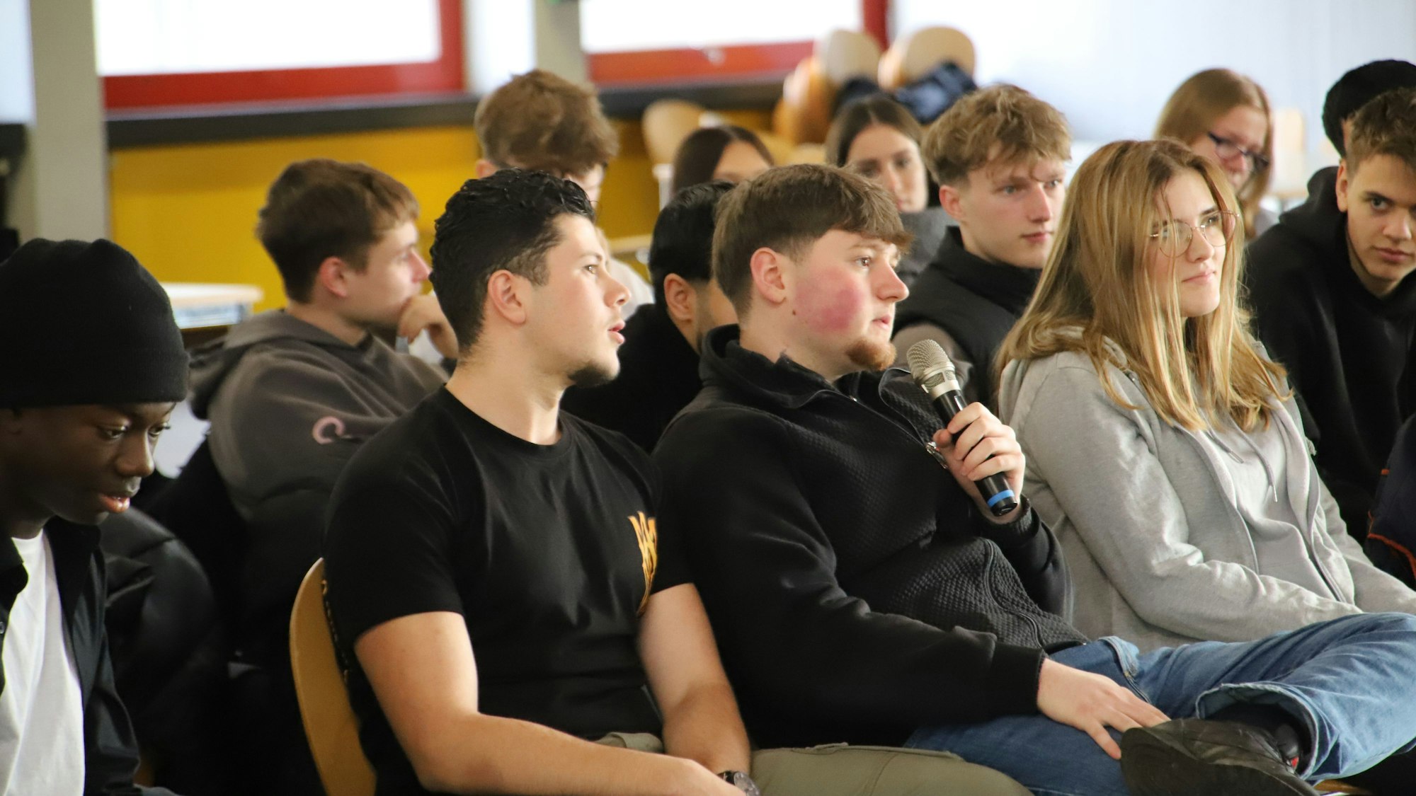 Schüler und Schülerinnen sitzen auf Stühlen, einer hat ein Mikrofon in der Hand.