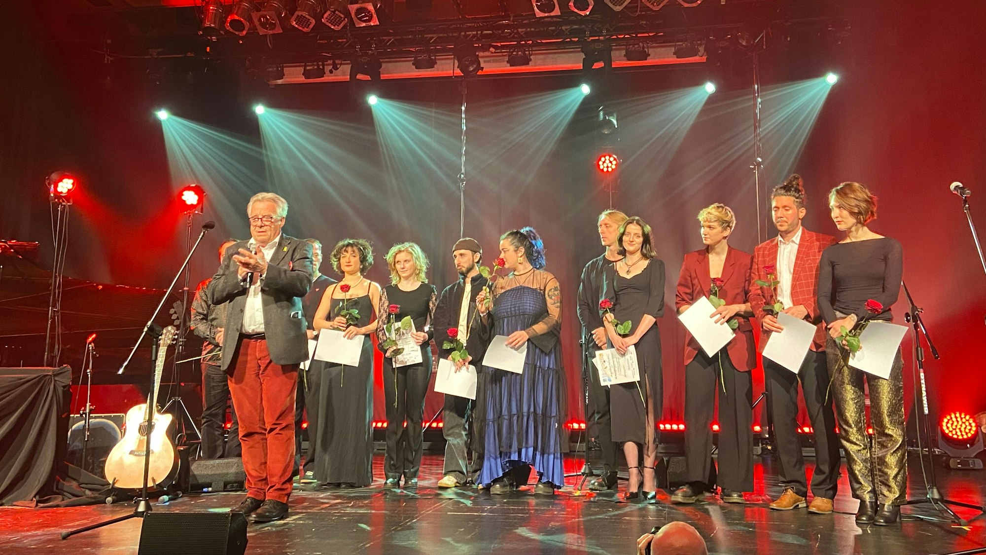 Blick auf eine Bühne, auf der Teilnehmer eines Chanson-Wettbewerbs stehen, sie haben alle eine Rose in der Hand.